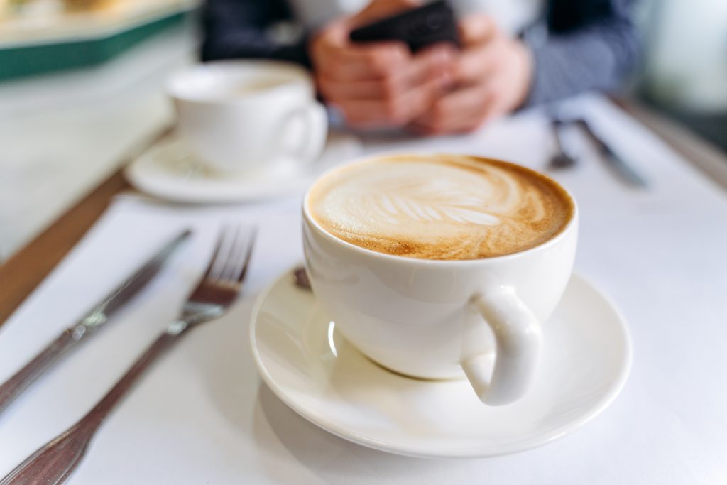 White cup of coffee on the table in a coffee shop. Man using a phone on the background